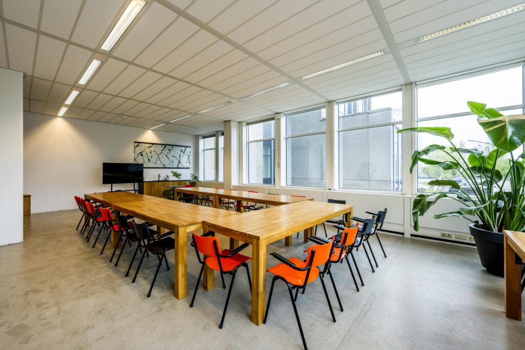 Modern meeting room at Laan van Kronenburg with wooden tables, colorful chairs, large windows, and indoor plants.